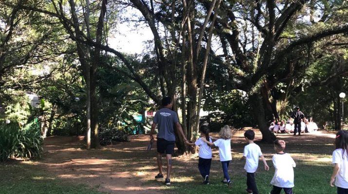 na imagem há uma grande praça com crianças em fila de costas e um educador vestindo cinza na frente