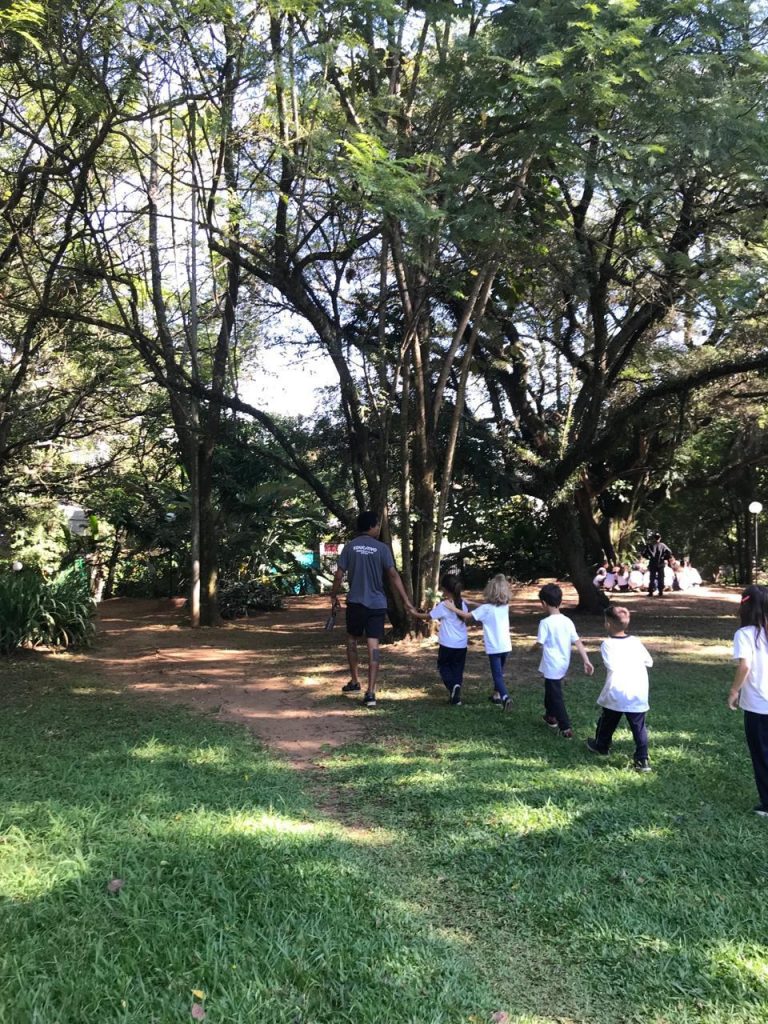 na imagem há uma grande praça com crianças em fila de costas e um educador vestindo cinza na frente